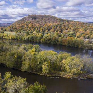 An aerial view of a river running through agricultural land, with wooded mountains and cloudy blue skies in the background.