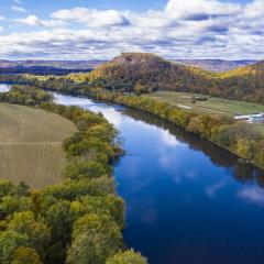 Aerial view with a river in the center buffered by trees and agricultural land on both sides. In the background there are forested mountains with fall foliage. There are cloudy blue skies above.
