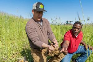 NRCS employee pointing to part of root in a rootball a farmer is holding. They are kneeling in a field of young wheat/